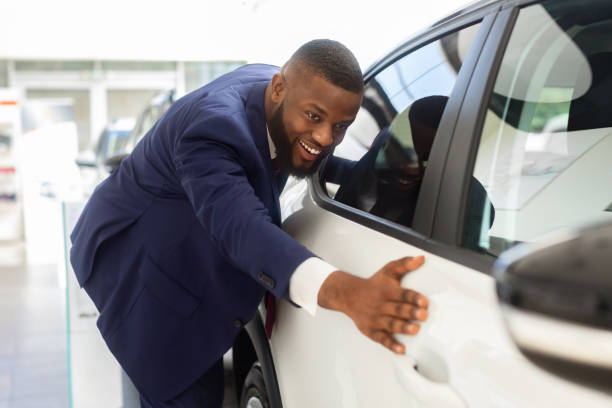 Happy Black Businessman In Suit Touching His New Car In Showroom After Buying, Excited African American Male Customer Admiring New Vehicle While Standing In Dealership Salon, Closeup Shot